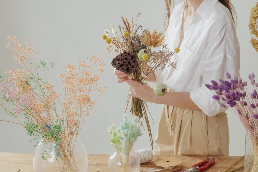 A woman arranges a bouquet of dried flowers, including Eryngium, with colorful dried flowers and craft supplies on a wooden table nearby